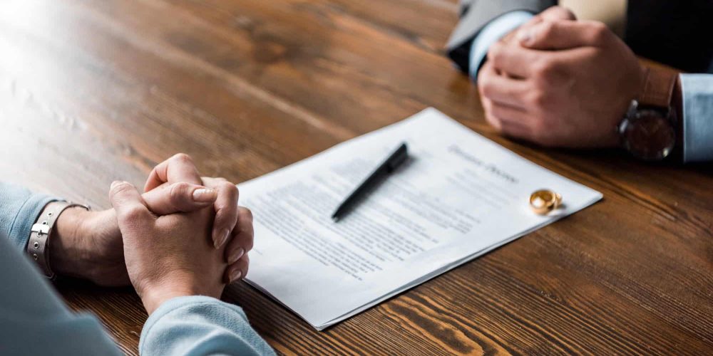 partial view of hands of lawyer and client, divorce decree and wedding rings on table