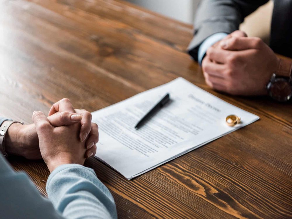partial view of hands of lawyer and client, divorce decree and wedding rings on table